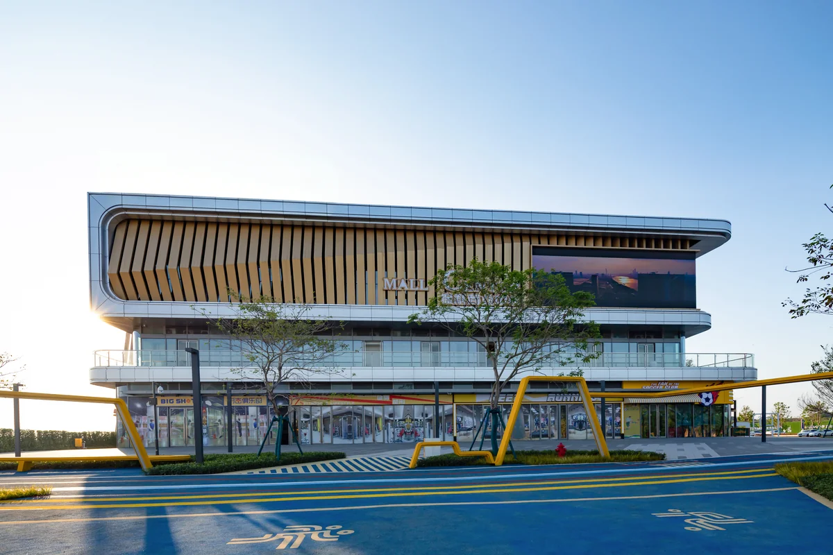 Completed two-storey commercial showroom in Bangkok Sukhumvit district, with a deep cantilevered overhang clad in warm wood-grain vertical aluminium louvers, large ground-floor glazing and a black LED display panel